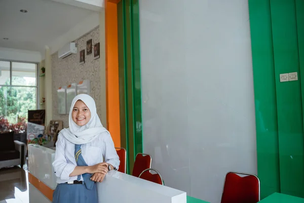 a smiling Indonesian high school student wearing a headscarf leaning back in front of the school