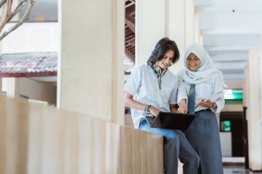 İki lise öğrencisi dizüstü bilgisayar kullanarak ve kitap taşıyarak gülümsüyor.