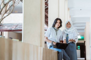 İki lise öğrencisi bir dizüstü bilgisayar kullanarak ve kitap taşıyarak kameraya gülümsüyor.