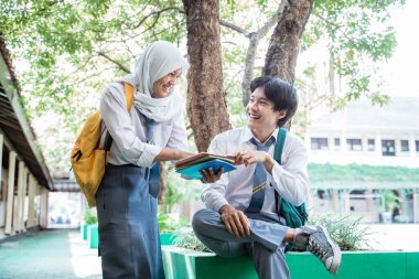 Gülümseyen bir oğlan ve peçeli bir kız Endonezya lise üniforması içinde ağacın altında kitap okurken sohbet ediyorlar.