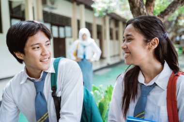 Yakışıklı bir oğlan ve liseli güzel bir kız, arka planda kıskanç bir jestle peçeli bir kız dururken sohbet ediyorlar.