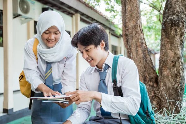 Lise üniformalı bir oğlan ve peçeli bir kız birlikte kitap okurken ağacın altında sohbet ediyorlar.
