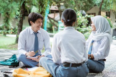 Lise üniformalı üç genç yerde oturan grup görevlerini tartışıyor.