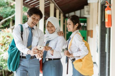 Bir erkek ve bir arkadaşının lise öğrencileri sohbet ederken cep telefonu ekranını işaret ederek birlikte bakarlar.