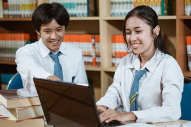 İki lise öğrencisi masada dizüstü bilgisayarla ders çalışarak sohbet ediyorlar.