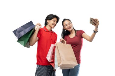 Alışveriş torbaları taşıyan Asyalı genç bir çift fotoğraf çektirirken telefonda poz veriyor.