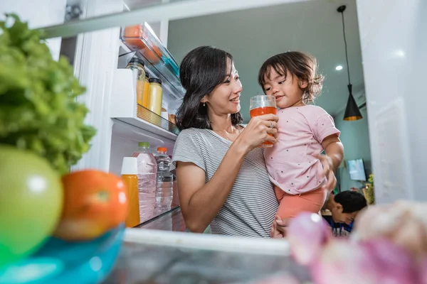 mother carrying her daughter while taking a glass of juice inside the fridge