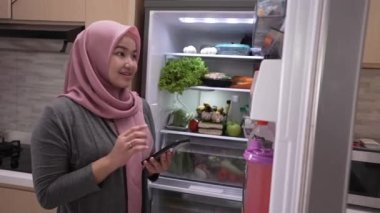 Müslüman genç bir kadın buzdolabını açarken akıllı telefonunu market alışverişi için kullanıyor.