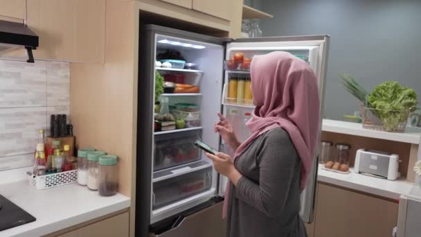 Mujer joven musulmana usando su teléfono inteligente para comprar comestibles mientras abre su nevera — Vídeos de Stock