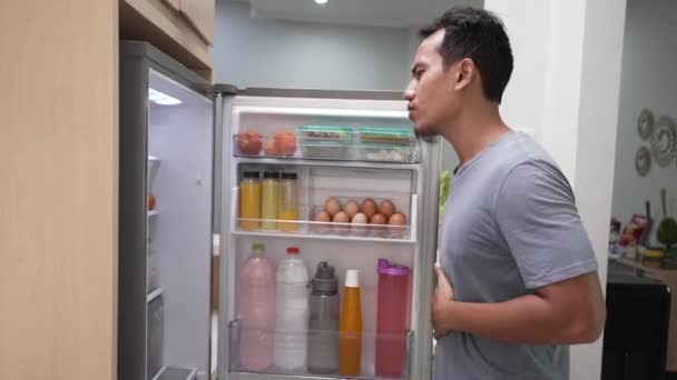 Sultende asiatisk mann holder seg i magen mens han åpner kjøleskapsdøra og ser etter noe å spise – stockvideo