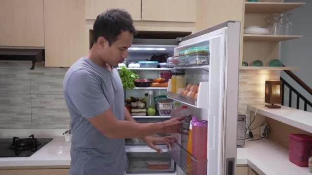 Жаждущий молодой человек берет напиток в холодильнике и пить его, пока холодильник еще открыт — стоковое видео
