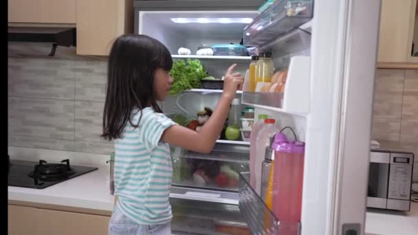 Ung asiatisk flicka öppna kylskåpsdörren letar efter mat och äta frukt — Stockvideo