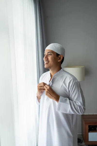 Pria muslim asia berdiri di dekat jendela berpakaian sebelum pergi ke masjid — Stok Foto