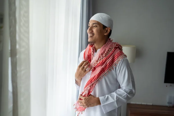 Pria muslim asia berdiri di dekat jendela berpakaian sebelum pergi ke masjid — Stok Foto