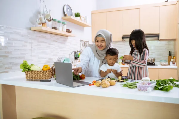 Ibu muslim menonton video memasak di laptop dan membuat makan malam dengan dua anaknya — Stok Foto