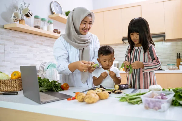 Ibu muslim menonton video memasak di laptop dan membuat makan malam dengan dua anaknya — Stok Foto