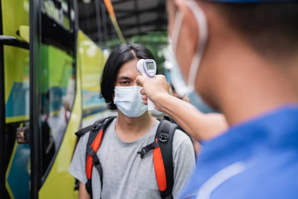 Close up de uma tripulação de ônibus em uniformes azuis e um chapéu usando uma arma térmica inspeciona o passageiro masculino na máscara — Fotografia de Stock