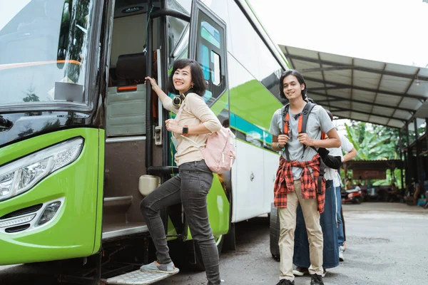 Asian woman carrying a backpack and headphones while holding the door handle gets into the bus with the background of passengers lining