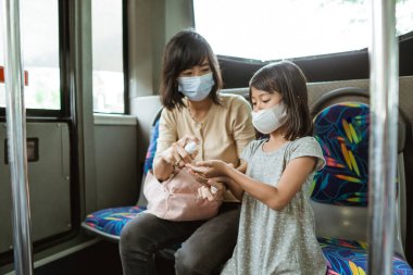 Bir kadın ve maske takan küçük bir kız el dezenfektanı kullanarak bankta oturuyorlar.