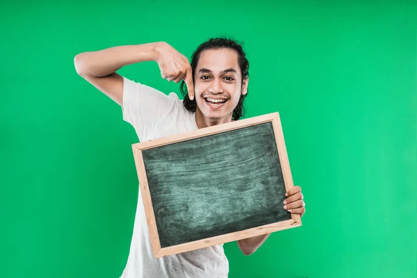 Junger Mann mit langen schwarzen Haaren hält und zeigt auf etwas auf einer leeren Tafel mit lächelndem Glück — Stockfoto