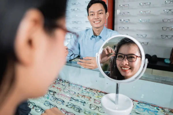 Una donna che indossa occhiali e che riflette in un bicchiere sullo sfondo di una vetrina per occhiali e un dipendente — Foto Stock