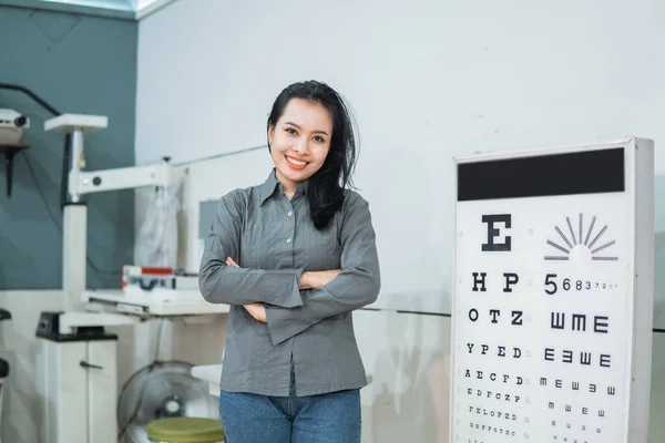 Жінка-лікар позує поруч з набором тестів для очей, розташованим в екзаменаційній кімнаті Стокове Зображення