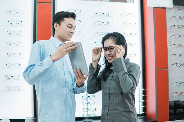 Un asistente de tienda sosteniendo una tableta mientras una cliente se prueba las gafas para mirar la pantalla de la tableta mientras está de pie —  Fotos de Stock