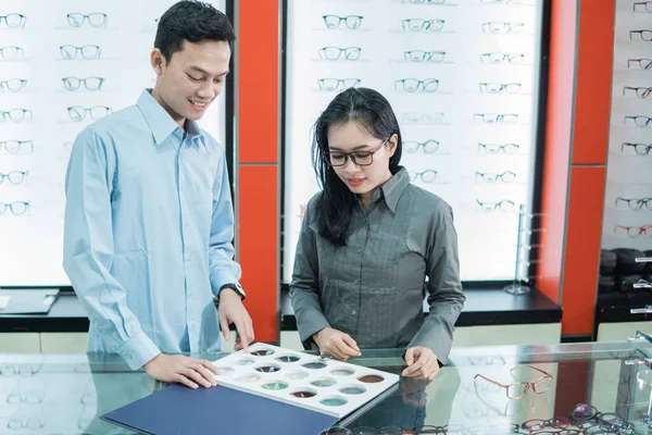 Un empleado de tienda masculino sostiene una muestra de lentes de gafas y un cliente señala su dedo para seleccionar lentes de gafas —  Fotos de Stock