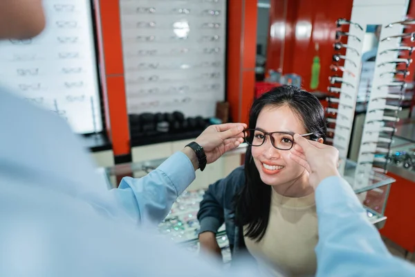 Una mano de los empleados está ayudando a ponerse un par de gafas que una mujer que ha hecho un examen ocular ha elegido —  Fotos de Stock