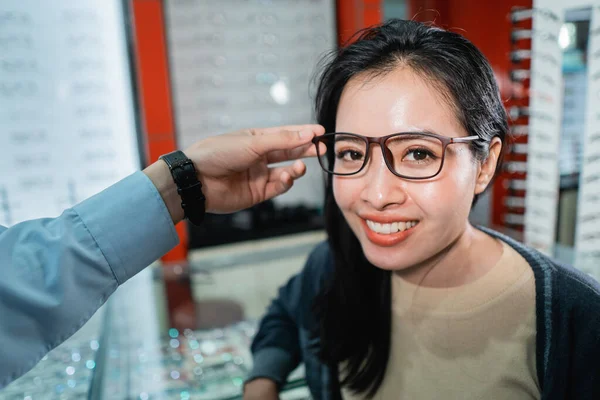 Una mano de los empleados está ayudando a ponerse un par de gafas que una mujer que ha hecho un examen ocular ha elegido —  Fotos de Stock