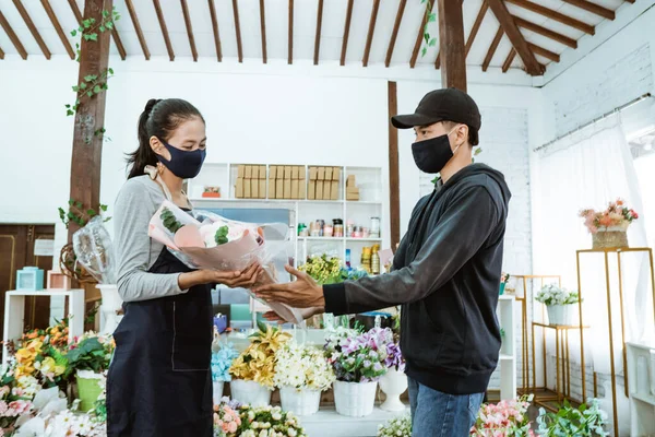 Smiling young female shopkeeper wearing a face mask and apron. servicing male flannel flower buyers — Stock Photo, Image