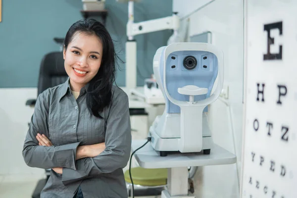 Seorang dokter perempuan berpose di samping alat tes mata terletak di ruang pemeriksaan Stok Foto Bebas Royalti