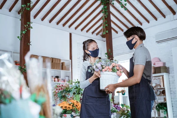 Junges Paar Blumenhändler mit Schürze und Gesichtsmaske hält Eimerblume lächelnd einander helfend — Stockfoto