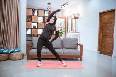 Spor kıyafetleri giymiş genç bir kadın vücudunu ısıtıyor. Bir eli belinde, bir eli yukarıda, yana doğru eğiliyor.