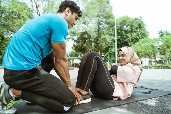 Um jovem ajuda a segurar as pernas de uma menina velada fazendo sit ups enquanto se exercita ao ar livre — Fotografia de Stock