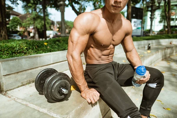 Közeli izmok alakítani egy izmos férfi, amikor ül kezében egy ivópoharat közelében a súlyzók — Stock Fotó