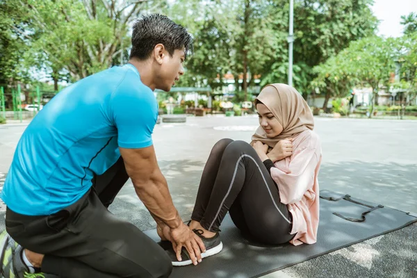 若い男は、屋外スポーツ中にABSワークアウトを行うベールの少女の足を保持するのに役立ちます — ストック写真