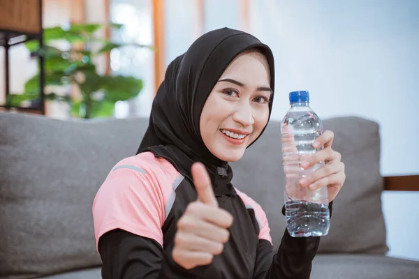 Крупным планом азиатская женщина в вуали спортивная улыбка, держа бутылку с большим пальцем вверх, сидя на полу — стоковое фото