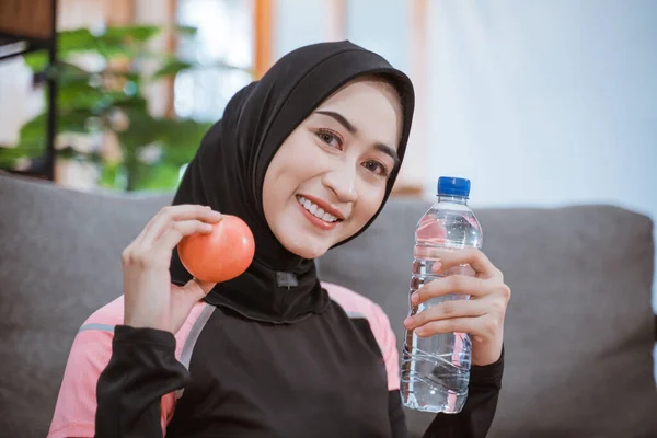Primer plano de una mujer asiática en un velo deportivo sonríe mientras sostiene una botella de bebida con gesto de mano ofrece una manzana mientras está sentado en el suelo —  Fotos de Stock