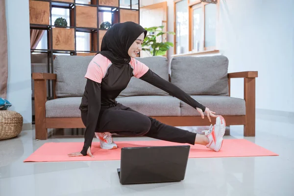 Une femme portant un hijab vêtements de gymnastique lors de l'accroupissement s'étend avec une jambe tirée latéralement et tenu d'une main lorsqu'il est devant un ordinateur portable — Photo