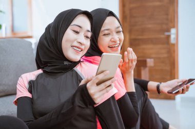 Spor kıyafetleri giyen iki kız yerde otururken cep telefonunun ekranına bakıp gülüyorlar.