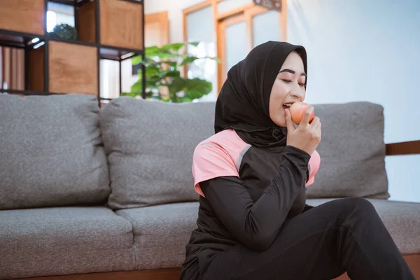Una mujer asiática en un velo ropa deportiva mientras sostiene y muerde una manzana con un sofá de fondo en casa —  Fotos de Stock