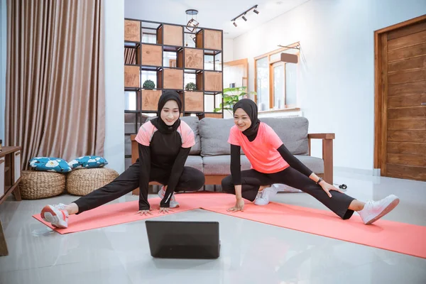 Deux jeunes femmes portant un hijab vêtements de gymnastique accroupi s'étend avec une jambe tirée latéralement tandis qu'en face d'un ordinateur portable — Photo