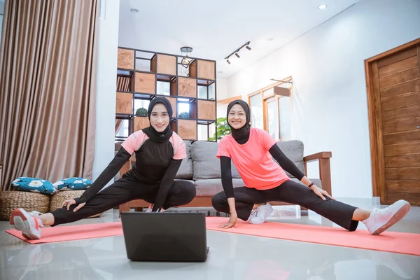 Deux jeunes femmes portant un hijab vêtements de gymnastique sourit à la caméra quand accroupi étire avec une jambe tirée latéralement tandis qu'en face d'un ordinateur portable — Photo