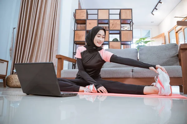 Una mujer usa un traje de gimnasio de hijab cuando está sentada se estira con una pierna hacia un lado y se sostiene con una mano — Foto de Stock