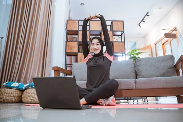 Une femme dans un hijab vêtements de sport s'assoit jambes croisées tout en s'étirant les mains vers le haut tout en regardant un ordinateur portable — Photo