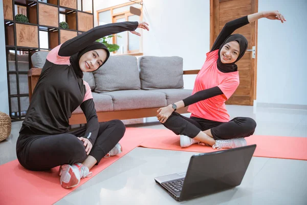 Dos mujeres que usan ropa deportiva de hijab se sientan con las piernas cruzadas en el suelo con sus cuerpos inclinados hacia un lado y las manos arriba mientras se calientan — Foto de Stock