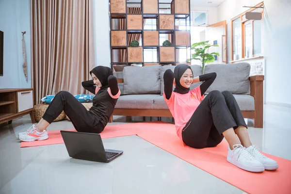 Dos mujeres en ropa deportiva hijab haciendo sentadas para entrenar los músculos abdominales en frente de una computadora portátil — Foto de Stock