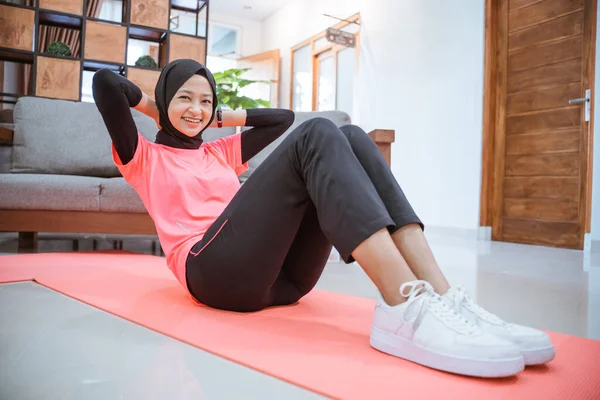 Ein Mädchen im Hijab-Turnoutfit lächelt, während es ein Sit-up auf einer Matte auf dem Boden macht — Stockfoto