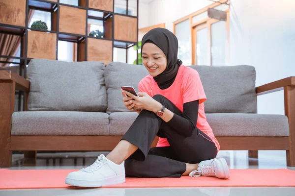 Aziatisch meisje in hijab sportkleding glimlacht kijken naar een mobiele telefoon terwijl zitten op de vloer — Stockfoto